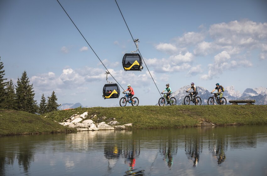 Vier Mountainbiker fahren neben einem Teich unter zwei Planai-Gondeln mit Blick auf Berge und bewaldete Landschaft | © Planai-Hochwurzen-Bahnen GesmbH