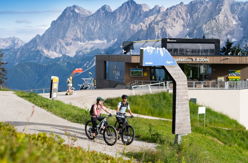 Start der Flowline am Bikepark Planai bei der Bergstation auf 1830 m | © Planai-Hochwurzen-Bahnen GesmbH Zwei Mountainbiker stehen am Flowline-Start vor der Planai-Bergstation auf 1830 m, mit dem Dachsteinmassiv im Hintergrund | © Planai-Hochwurzen-Bahnen GesmbH
