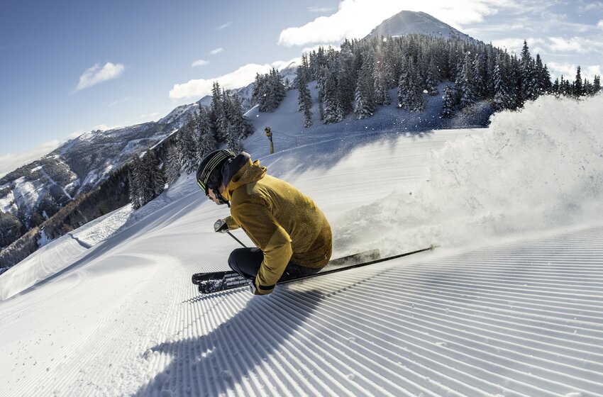 Ein Skifahrer zieht einen tiefen Carving-Schwung über eine frisch präparierte Piste vor verschneiten Bergen und Tannen. | © Mirja Geh