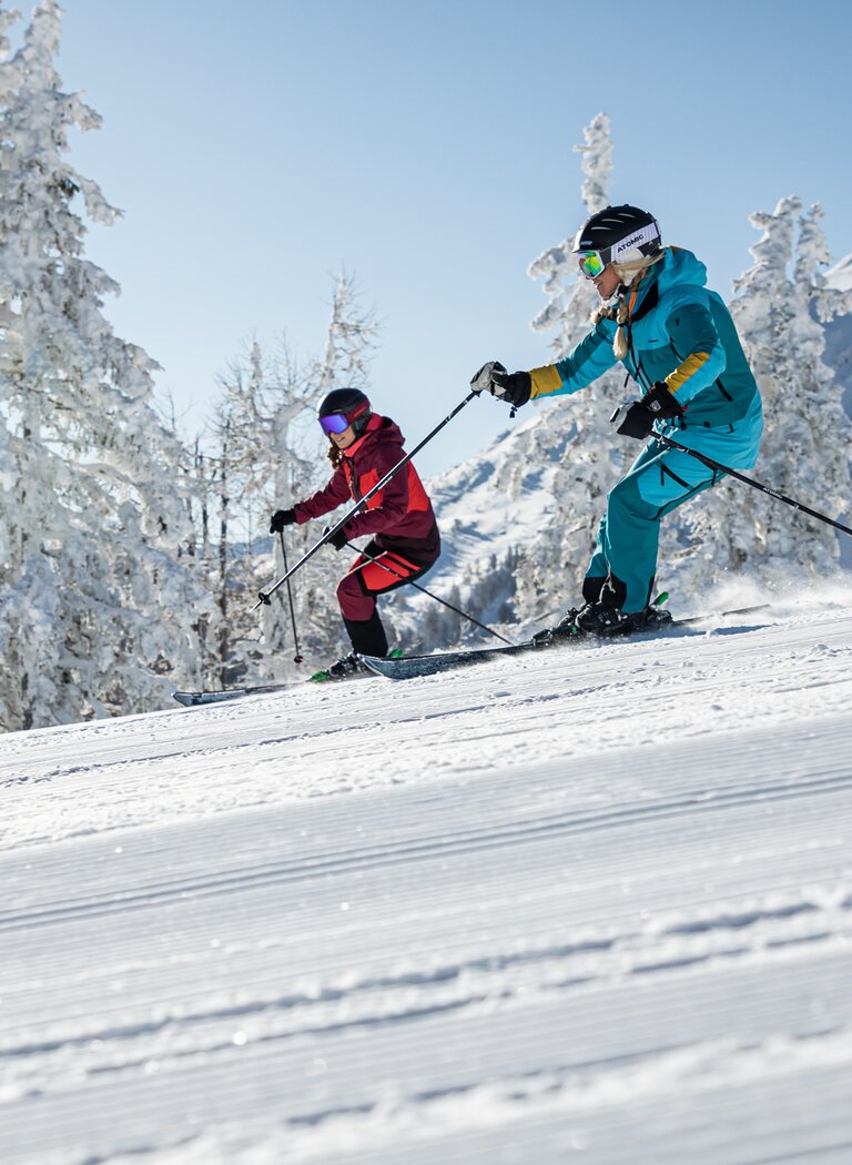 Zwei Skifahrerinnen fahren nebeneinander auf einer frisch präparierten Piste umgeben von verschneiten Bäumen und Bergen. | © Reiteralm Bergbahnen GmbH & Co.KG