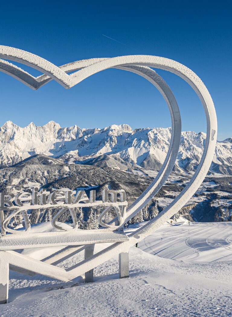 Herzplatz auf der Reiteralm mit Winterpanorama und Blick auf Dachstein | © Reiteralm Bergbahnen GmbH & Co.KG Verschneite Herz-Skulptur mit „Reiteralm“-Schriftzug vor winterlichem Dachstein-Panorama bei strahlend blauem Himmel. | © Reiteralm Bergbahnen GmbH & Co.KG