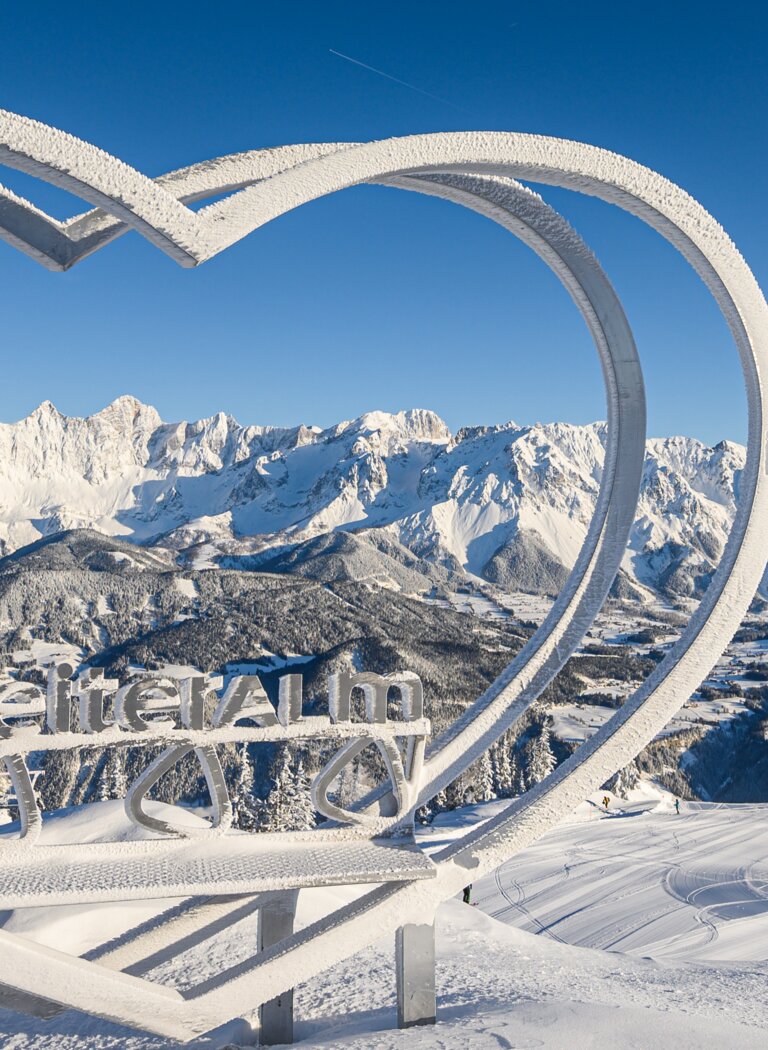 Snow-covered heart sculpture with “Reiteralm” sign and Dachstein panorama under a clear blue winter sky. | © Reiteralm Bergbahnen GmbH & Co.KG