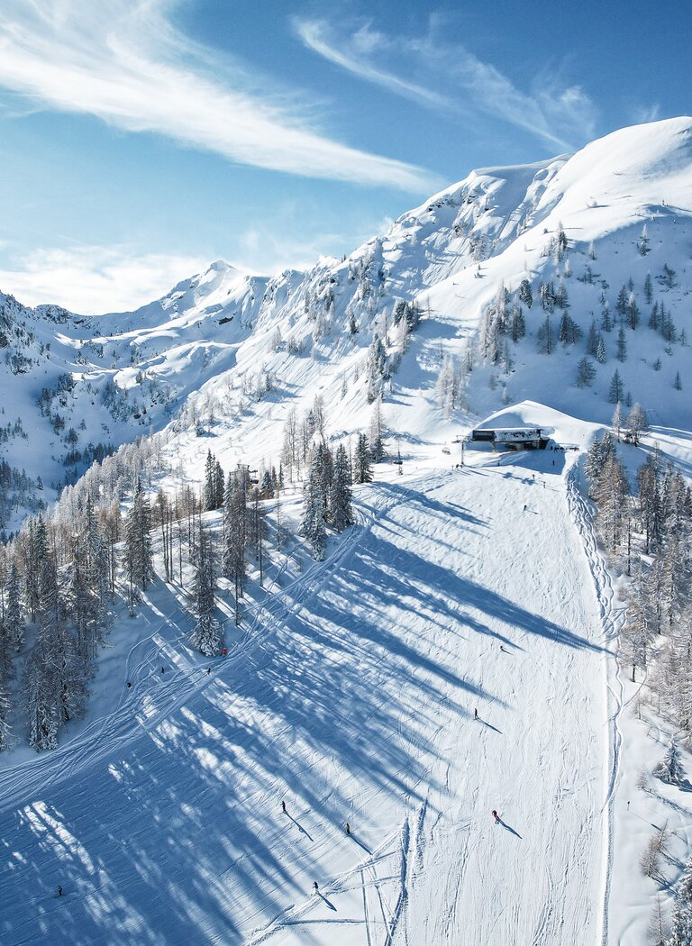 Snow-covered ski slope with several skiers, lined by trees and surrounded by high mountains under clear sky | © Reiteralm Bergbahnen GmbH & Co.KG