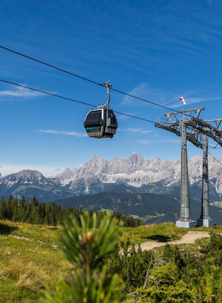 Two Reiteralm gondolas float above alpine meadow with wide view of Dachstein range under clear blue sky. | © Reiteralm Bergbahnen GmbH & Co.KG
