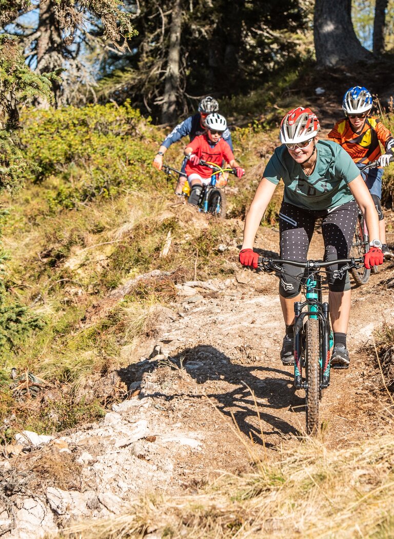 Group of adults and kids riding mountain bikes on narrow forest trail through sunny alpine terrain. | © Reiteralm Bergbahnen GmbH & Co.KG