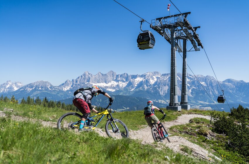 Two mountain bikers ride a winding trail beneath a gondola lift with snow-capped Alps and clear blue sky. | © Reiteralm Bergbahnen GmbH & Co.KG