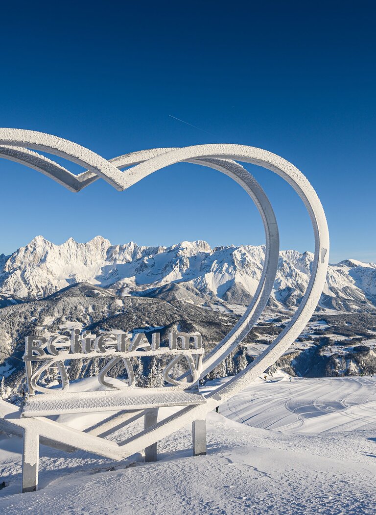 Snow-covered heart sculpture with Reiteralm logo and Dachstein range in the background under clear blue sky. | © Reiteralm Bergbahnen GmbH & Co.KG
