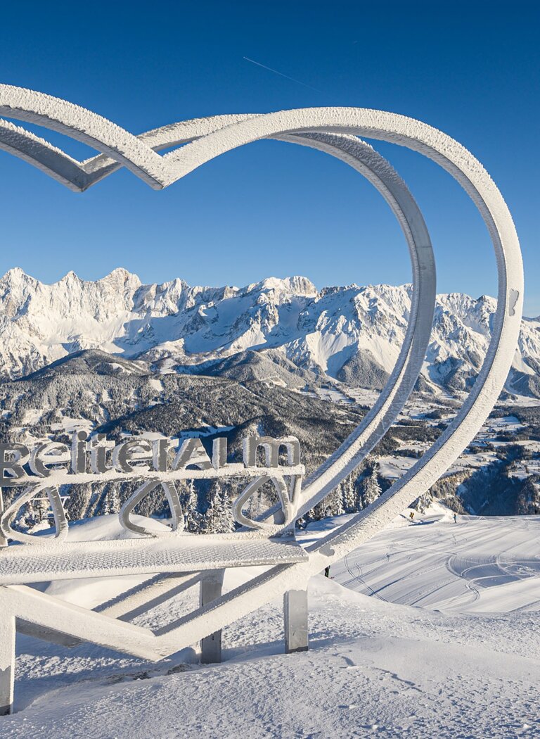 Snow-covered heart sculpture with Reiteralm logo and Dachstein range in the background under clear blue sky. | © Reiteralm Bergbahnen GmbH & Co.KG