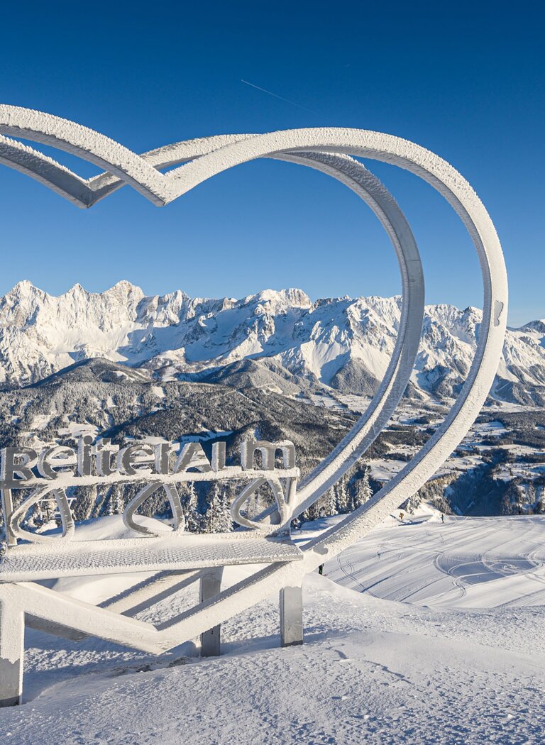 Verschneite Herzskulptur mit Reiteralm-Schriftzug vor Panorama des verschneiten Dachsteinmassivs unter blauem Himmel. | © Reiteralm Bergbahnen GmbH & Co.KG