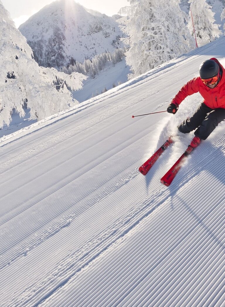 Skifahrer in roter Jacke fährt sportlich über frisch präparierte Piste, umgeben von verschneiten Bäumen im Sonnenschein. | © Reiteralm Bergbahnen GmbH & Co.KG