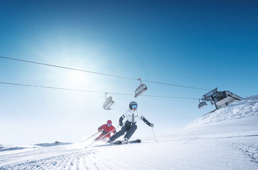 Zwei Skifahrer carven schwungvoll unter Sessellift im Sonnenschein | © Ski amadé GmbH Zwei Skifahrer carven auf frisch präparierter Piste unter Sessellift bei blauem Himmel und strahlender Morgensonne. | © Ski amadé GmbH